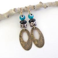 These handmade earrings feature gold brass hoops with a hand stamped texture accented with African batik bone beads and topped off with turquoise color evil eye glass and matte black glass beads.  Evil eye beads are protective amulets designed with concentric circles that mimic the appearance of a human eye. Beads with this symbolism have been used for thousands of years across different cultures to ward off bad luck, illness, or misfortune and push away the negative or envious energy of others.