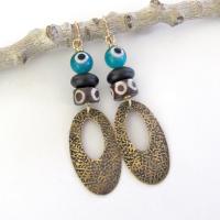 These handmade earrings feature gold brass hoops with a hand stamped texture accented with African batik bone beads and topped off with turquoise color evil eye glass and matte black glass beads.  Evil eye beads are protective amulets designed with concentric circles that mimic the appearance of a human eye. Beads with this symbolism have been used for thousands of years across different cultures to ward off bad luck, illness, or misfortune and push away the negative or envious energy of others.