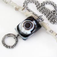 This handmade necklace features a natural eye agate stone accented with a hammered silver pewter circle. When cut at the right angle, this type of stone reveals a series of concentric rings that resembles the human eye. The stone in this piece has a bold matrix of black and white bands with a black circular center. It will make a great gift for a natural stone lover. 
