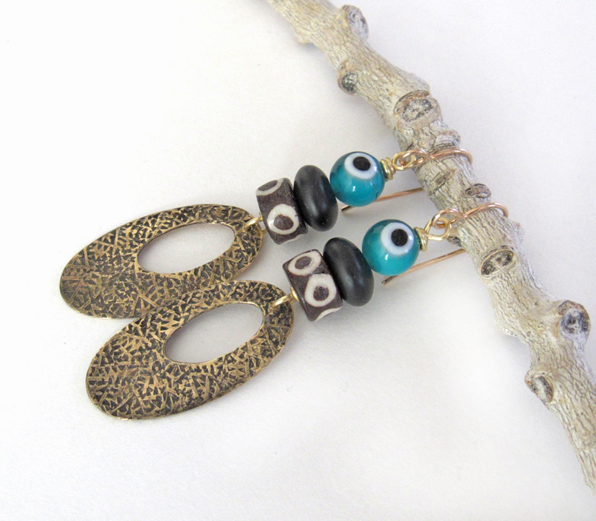 These handmade earrings feature gold brass hoops with a hand stamped texture accented with African batik bone beads and topped off with turquoise color evil eye glass and matte black glass beads.  Evil eye beads are protective amulets designed with concentric circles that mimic the appearance of a human eye. Beads with this symbolism have been used for thousands of years across different cultures to ward off bad luck, illness, or misfortune and push away the negative or envious energy of others.