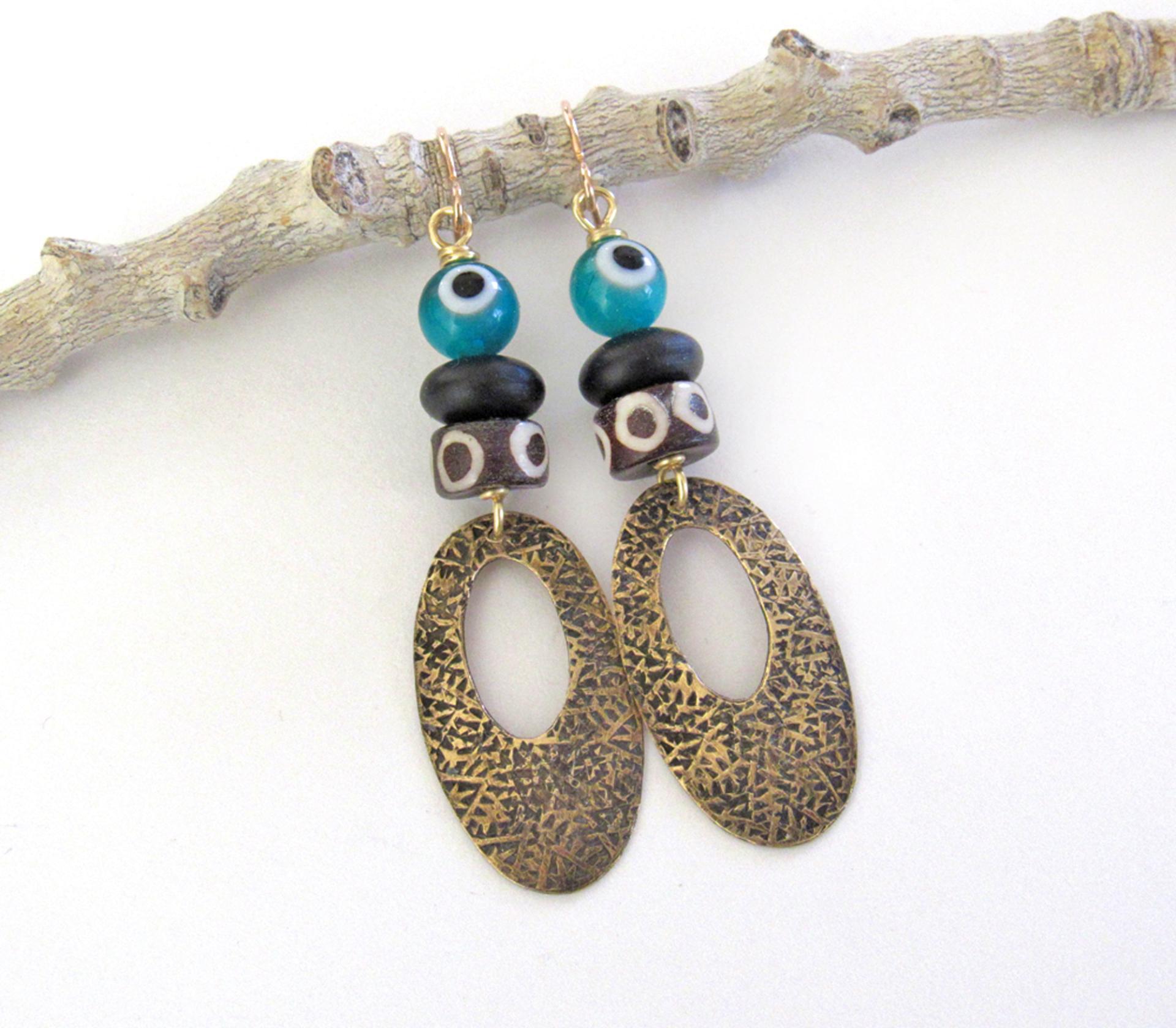 These handmade earrings feature gold brass hoops with a hand stamped texture accented with African batik bone beads and topped off with turquoise color evil eye glass and matte black glass beads.  Evil eye beads are protective amulets designed with concentric circles that mimic the appearance of a human eye. Beads with this symbolism have been used for thousands of years across different cultures to ward off bad luck, illness, or misfortune and push away the negative or envious energy of others.