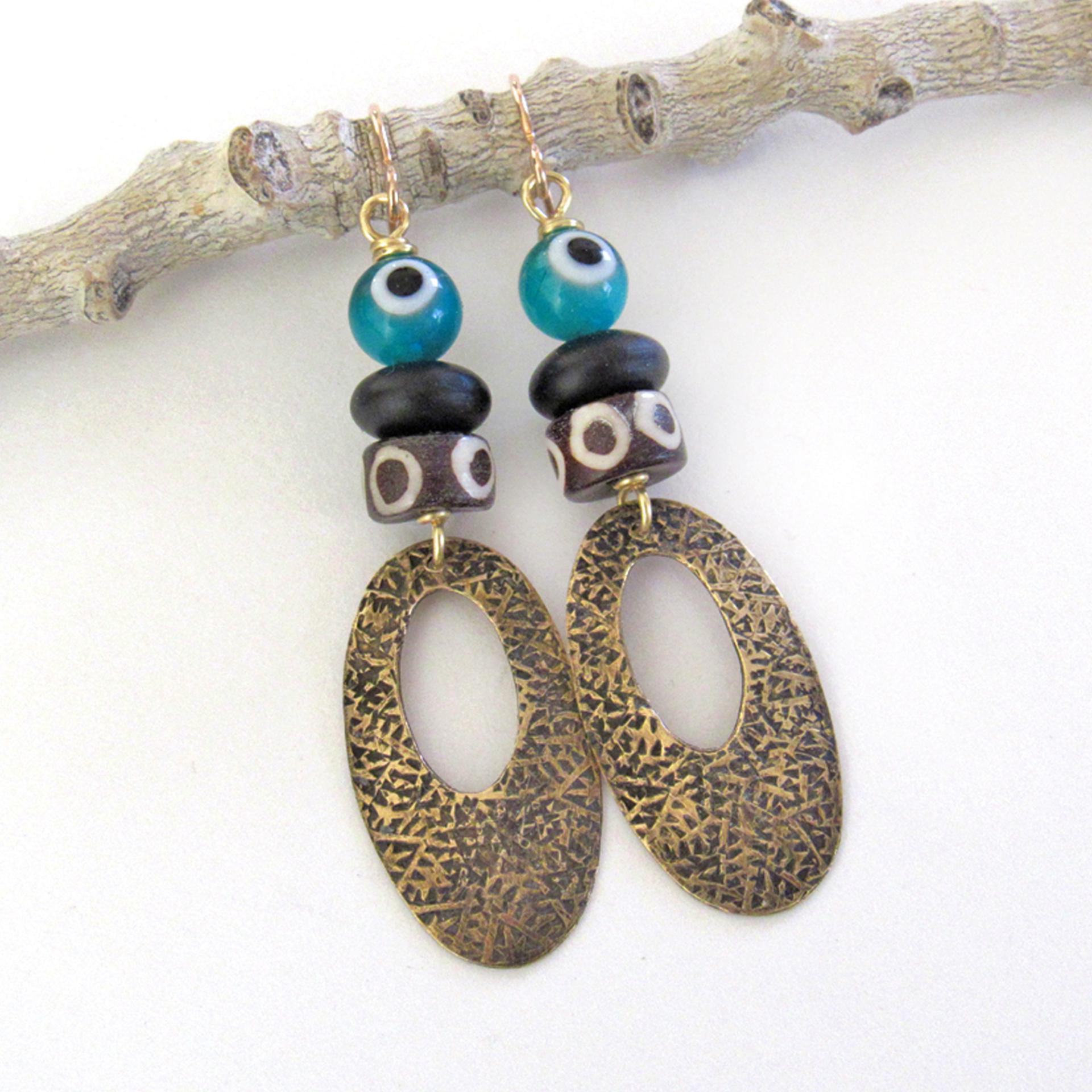 These handmade earrings feature gold brass hoops with a hand stamped texture accented with African batik bone beads and topped off with turquoise color evil eye glass and matte black glass beads.  Evil eye beads are protective amulets designed with concentric circles that mimic the appearance of a human eye. Beads with this symbolism have been used for thousands of years across different cultures to ward off bad luck, illness, or misfortune and push away the negative or envious energy of others.