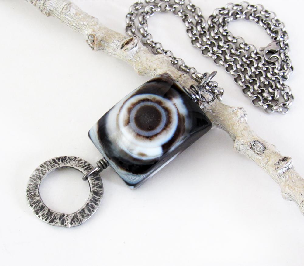 This handmade necklace features a natural eye agate stone accented with a hammered silver pewter circle. When cut at the right angle, this type of stone reveals a series of concentric rings that resembles the human eye. The stone in this piece has a bold matrix of black and white bands with a black circular center. It will make a great gift for a natural stone lover. 