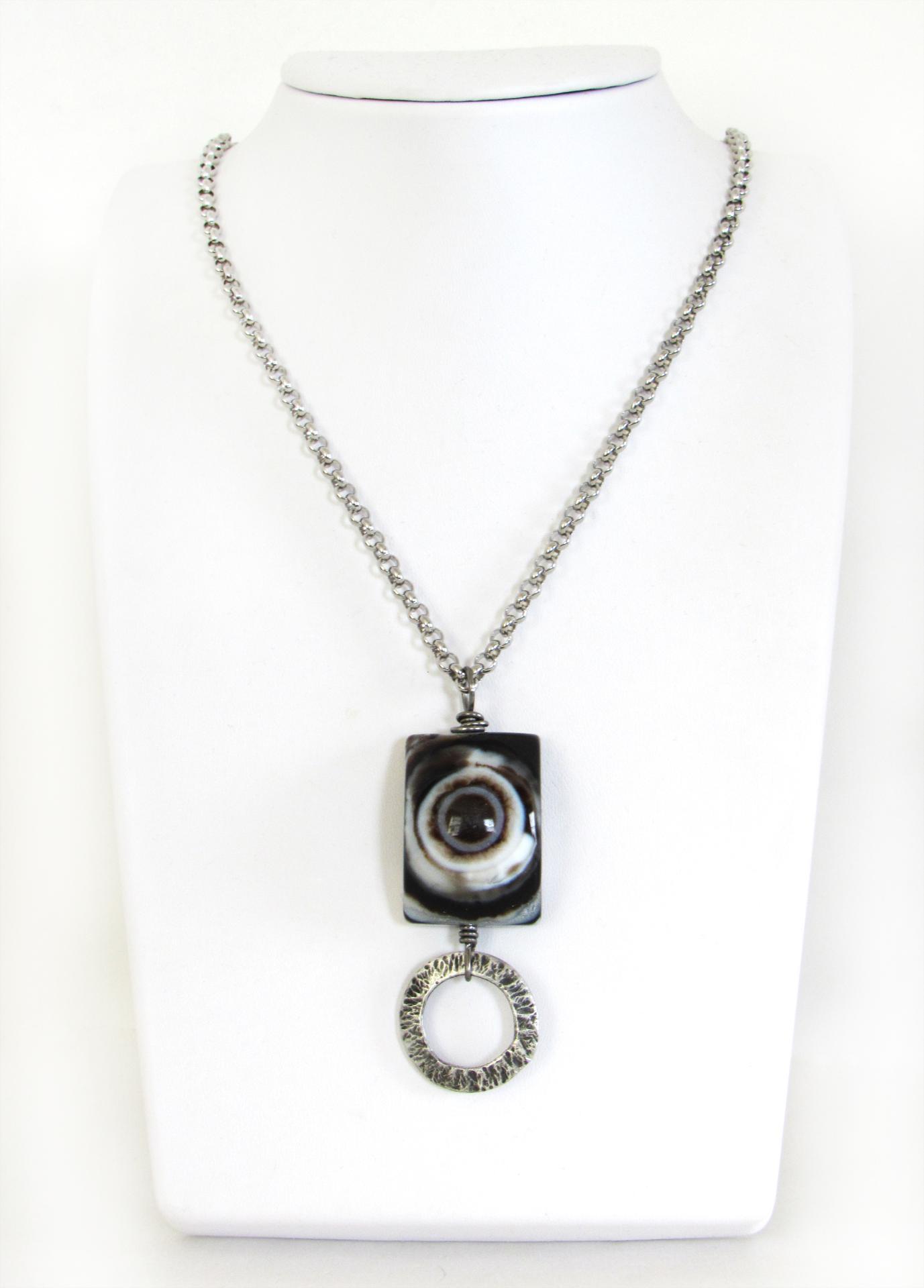 This handmade necklace features a natural eye agate stone accented with a hammered silver pewter circle. When cut at the right angle, this type of stone reveals a series of concentric rings that resembles the human eye. The stone in this piece has a bold matrix of black and white bands with a black circular center. It will make a great gift for a natural stone lover. 