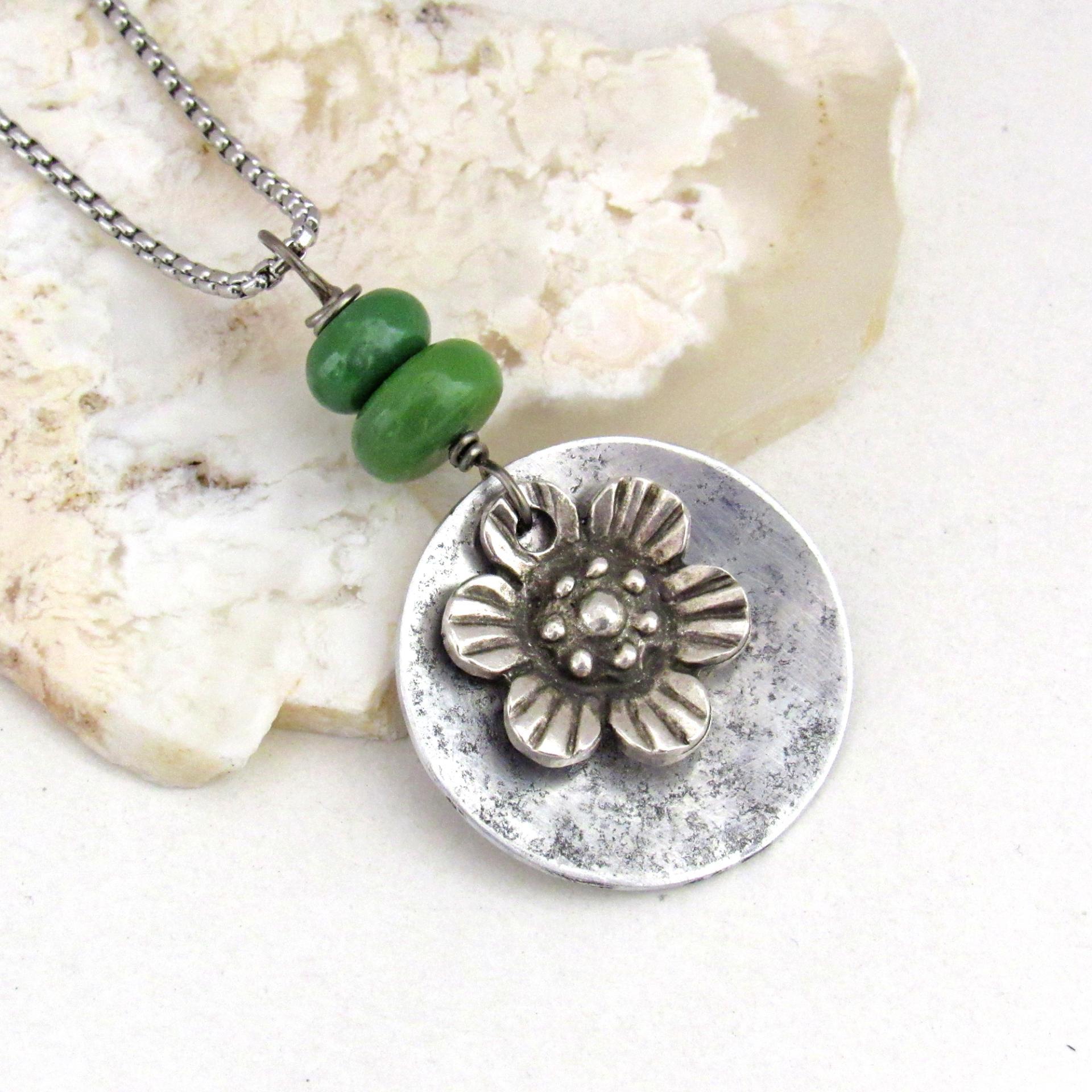 This handmade necklace features a silver tone pewter flower charm framed against an aluminum circle accented with a stack of natural green serpentine gemstones. It comes on a 20" stainless steel chain necklace with lobster claw closure. It'll make a great gift for anyone that loves flowers or nature.