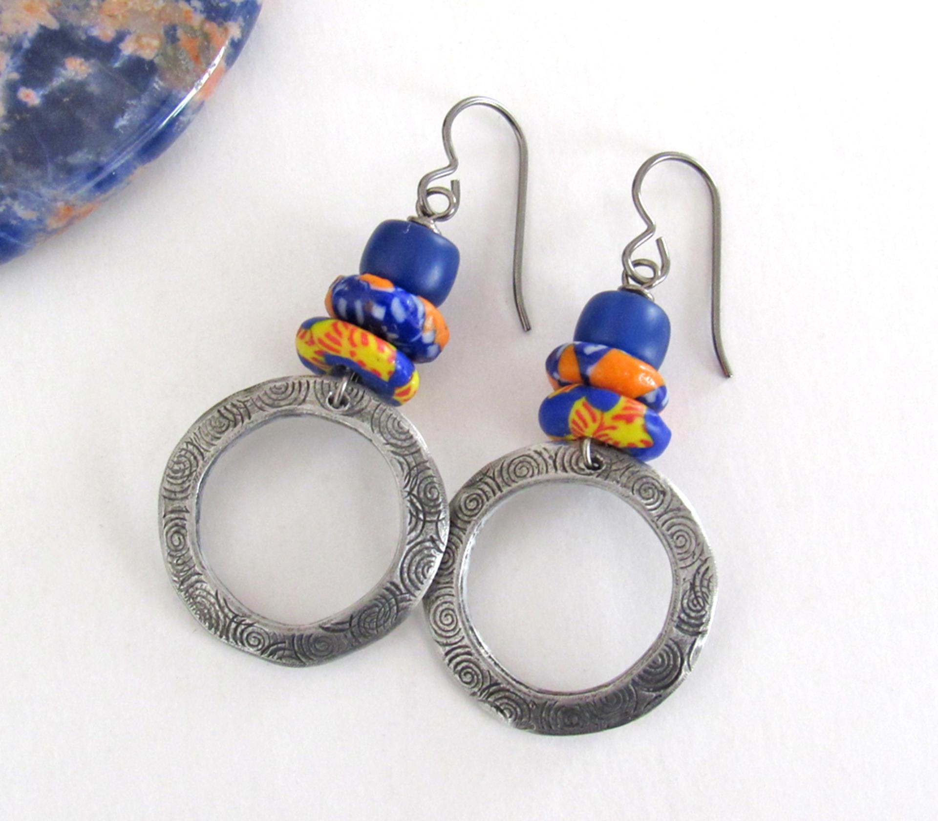 These handcrafted earrings feature silver pewter circle hoops with a hand stamped texture accented with colorful African glass beads and navy blue glass beads. They have an earthy, boho style and are lightweight and comfortable to wear.  MEASUREMENTS: Total length 2" from top of gray silver niobium (hypoallergenic) ear wires and 1” at widest point. The circles were created out of solid lead-free pewter; hand stamped and oxidized; and signed on the back with my initials.